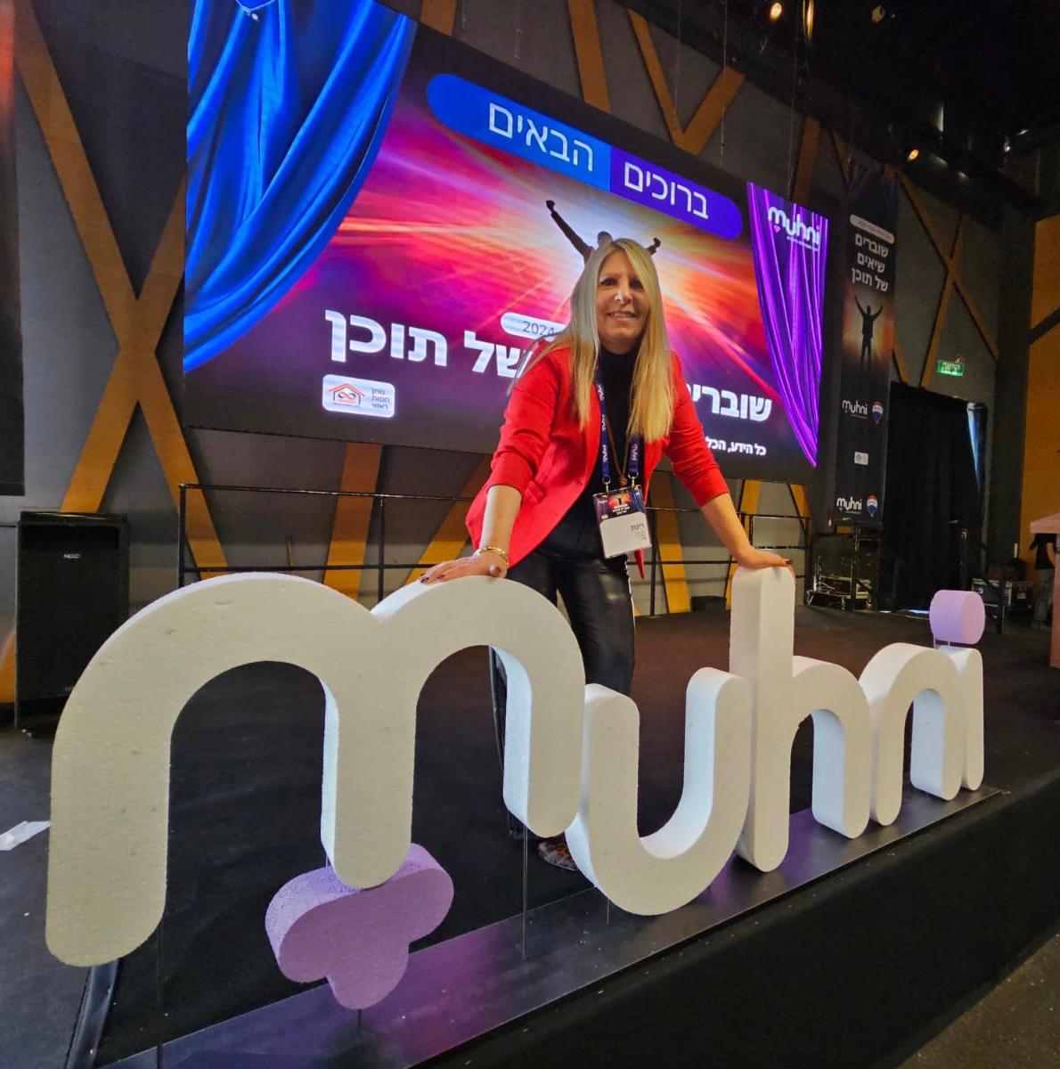רינת בכנס Muhni מול השלט הענק